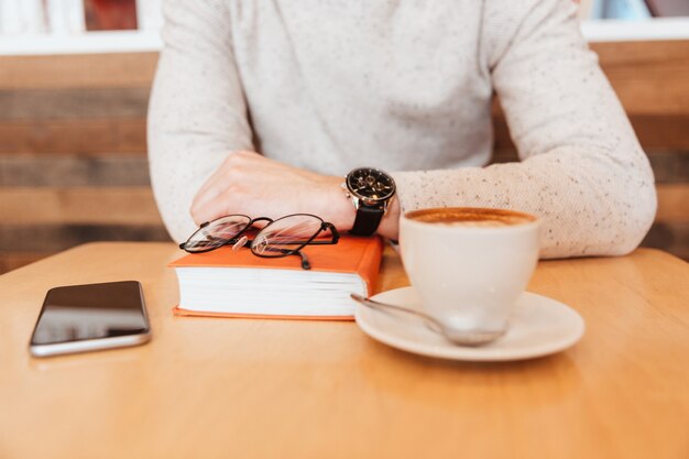 Abgeschnittenes Foto eines jungen Mannes im Hemd, der mit einer Tasse Kaffee und einem Buch im Café sitzt.