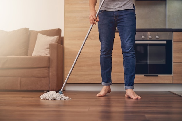 Abgeschnittenes Foto eines Mannes in Jeans und grauem T-Shirt, das Parkett mit speziellem Moping-Stick in der Wohnung putzt