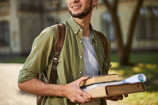 Abgeschnittenes Foto eines zufriedenen jungen Mannes mit dunklem Bart, der einen Stapel Bücher hält