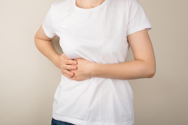 Abgeschnittenes Nahaufnahmefoto einer jungen Frau in weißem T-Shirt, die den rechten Oberbauch mit den Händen auf isoliertem grauem Hintergrund hält