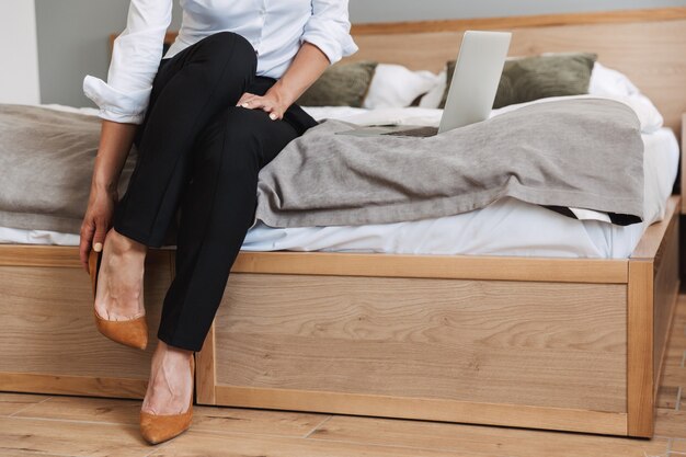 Abgeschnittenes Porträt einer kaukasischen erwachsenen Geschäftsfrau in formellem Anzug, die ihre Schuhe auszieht, während sie auf dem Bett sitzt und einen Laptop in der Wohnung benutzt