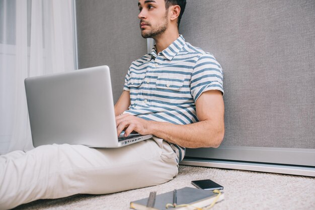 Abgeschnittenes Porträt eines fröhlichen jungen kaukasischen Mannes in stilvollem Hemd, der zu Hause auf dem Boden sitzt, mit offenem Laptop-PC auf seinem Schoß Hinweis auf Papier für Unternehmensgründung Menschen und Lifestyle-Konzept