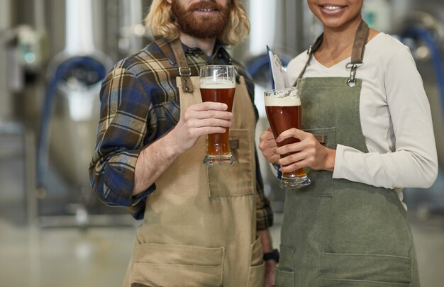 Foto abgeschnittenes porträt von zwei lächelnden jungen arbeitern, die bierglas halten und in die kamera schauen, während sie in der werkstatt in der brauerei stehen, kopierraum