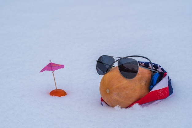 Abóbora crua engraçada com óculos e bandana americana em uma cama de neve e fundo branco, close-up. Ainda vida engraçada de natal quando você não pode ir de férias