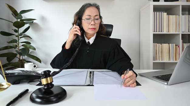 Abogada madura con uniforme de túnica sentada frente a una laptop y hablando por teléfono con sus clientes para brindar consultas legales