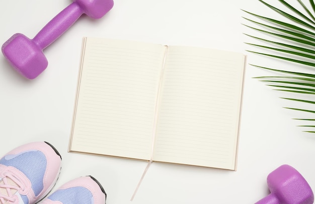 Abra el cuaderno en blanco, un par de zapatillas deportivas y pesas sobre un fondo blanco Lugar para grabar la vista superior