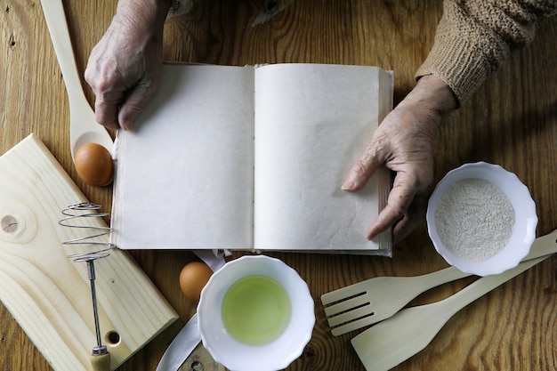 Abra o livro de receitas nas mãos de uma senhora idosa em frente a uma mesa com utensílios