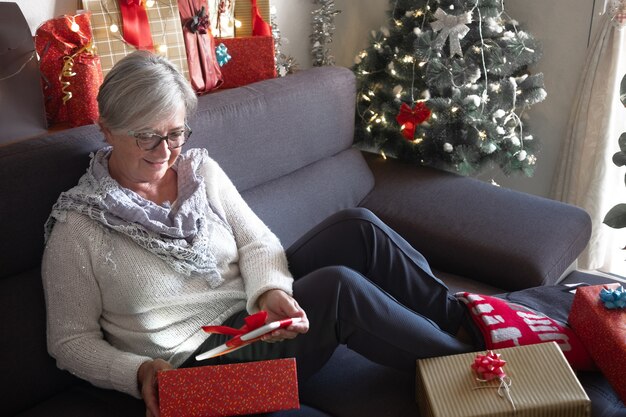 Abra uma caixa de presente e encontre um novo tablet branco como presente de Natal. Uma velha senhora de cabelos grisalhos sorri surpresa, sentada no canto do sofá. Árvore de natal e presentes em torno dela