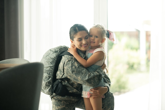 Abraçando garota adorável. mulher militar madura usando uma mochila e abraçando sua linda garota