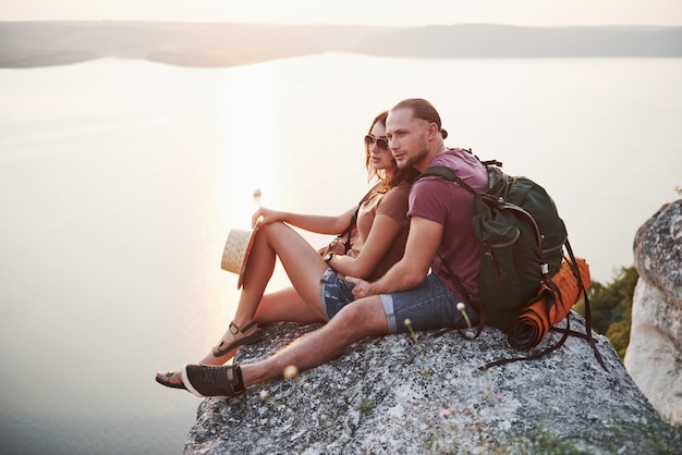 Abraçando o casal com mochila, sentado no topo da montanha de pedra, apreciando a vista costa um rio ou lago. Viajar ao longo de montanhas e costa, liberdade e conceito de estilo de vida ativo