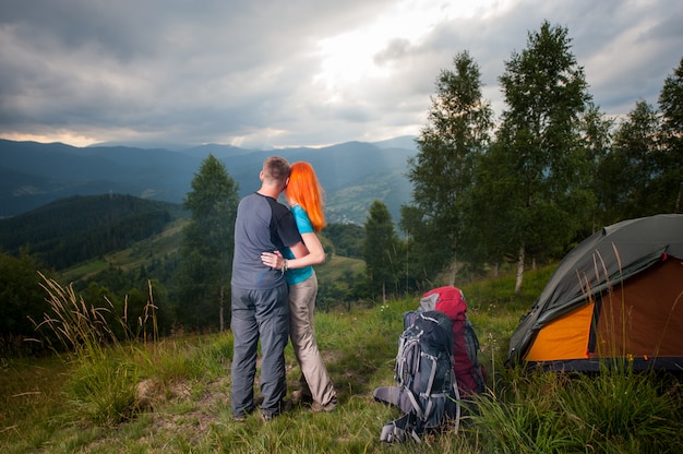 Abraçando o casal em pé de volta perto do acampamento e olhando para a distância nas montanhas, florestas e perfurando os raios do sol através de um céu nublado ao pôr do sol