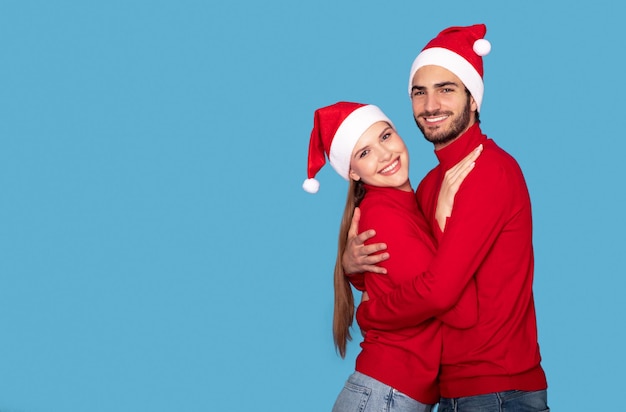 Abraçando o jovem casal com chapéus de Papai Noel posando sobre fundo azul no estúdio