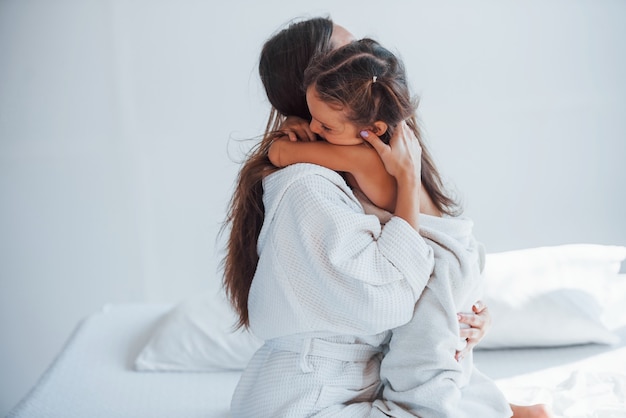 Abraçando-se. Jovem mãe com sua filha tem dia de beleza dentro de casa, no quarto branco.