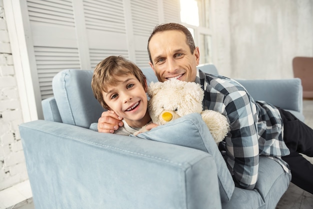 Abraçando. Um rapaz bonito e alegre, de cabelos louros, sorrindo e sentado no sofá com seu brinquedo e seu pai o abraçando