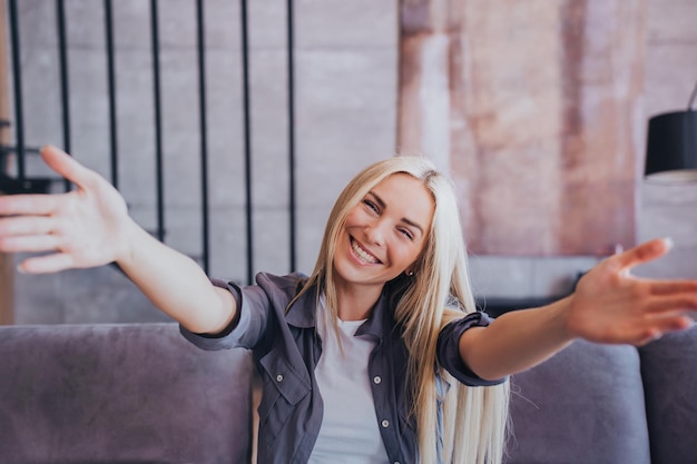 Abrace-me Linda loira Caucasiana em casual abre as mãos sorrindo largamente dá as boas-vindas ao abraço estando de bom humor Mulher muito alegre sentada no sofá aconchegante em casa Pessoas verdadeiras emoções