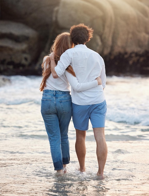 Abraço nas costas e casal andando na praia e tempo de qualidade nas férias de verão, fim de semana e romance Homem e mulher românticos abraçam amor e felicidade para viagens de férias e aventuras juntos