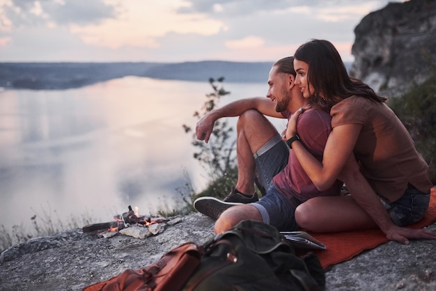 Abrazando pareja con mochila sentado cerca del fuego en la cima de la montaña disfrutando de la vista de la costa de un río o lago. concepto de libertad y estilo de vida activo