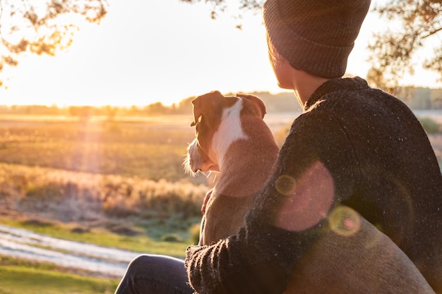Abrazando a un perro en la hermosa naturaleza al atardecer. Mujer frente al sol vespertino se sienta con su mascota junto a ella y disfruta de la belleza de la naturaleza
