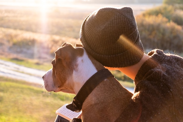 Abrazando a un perro en la hermosa naturaleza al atardecer. Mujer frente al sol vespertino se sienta con su mascota junto a ella