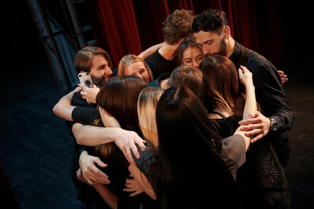 Abrazándose unos a otros Grupo de actores en ropa de color oscuro en el ensayo en el teatro