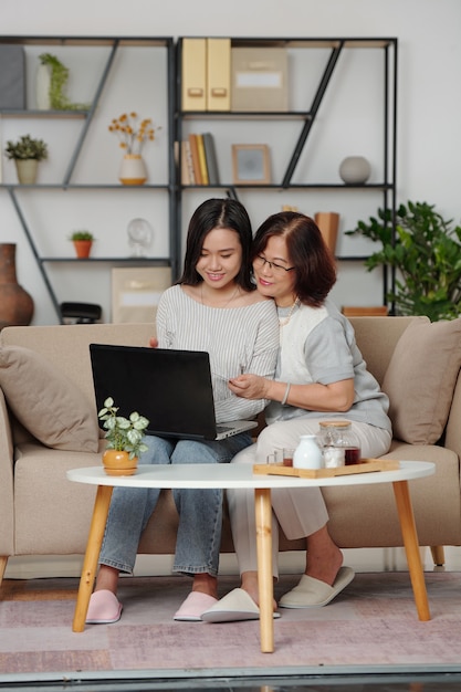 Abrazar a la feliz madre e hija asiáticas sentadas en el sofá con el portátil y hacer compras en la tienda online