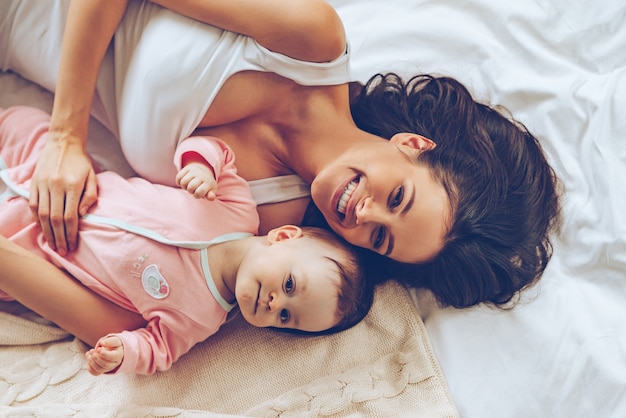 Abrazos matutinos. Vista superior de la alegre hermosa joven mirando a la cámara con una sonrisa mientras está acostado en la cama con su bebé