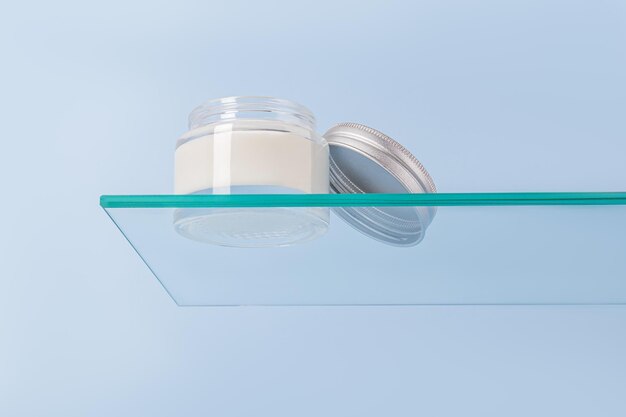 Abre un frasco de cosméticos transparente con crema blanca para el cuidado diario de la piel de la cara y el cuerpo en una superficie flotante de vidrio sobre un fondo azul