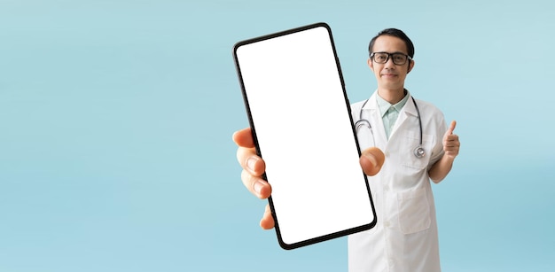 Foto abrigo de médico asiático con mano sosteniendo un gran teléfono inteligente aislado en fondo azul con caminos de recorte para el trabajo de diseño espacio libre vacío