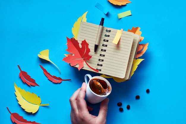 Abrir cuaderno en blanco con hojas de otoño y mano con café en azul