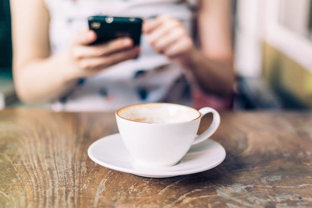 Foto abschluss herauf kaffeetasse mit der frau, die smartphone, weinleseton spielt.