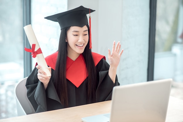 Abschluss mit einem Diplom. Netter asiatischer lächelnder Absolvent, der ihr Diplom hält und einen Videoanruf hat