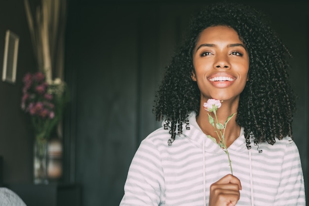 Abschluss oben einer hübschen schwarzen Frau mit dem gelockten Haar lächelnd mit einer Rosenblume sitzen auf dem Bett, das weg schaut