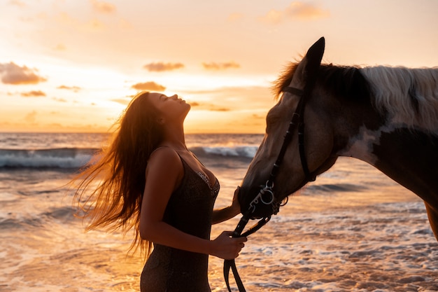 Abschluss oben: Junge Brunetteschönheit, die Spaß mit Pferd auf dem Strand hat. Sonnenuntergang