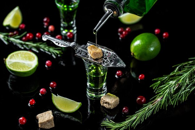 Absinth-Shots mit Zuckerwürfeln Absinth in eine Glasflasche Absinth mit braunen Zuckerpreiselbeeren und Limetten-Edelstahllöffel isoliert auf schwarzem Hintergrund Platz für Text