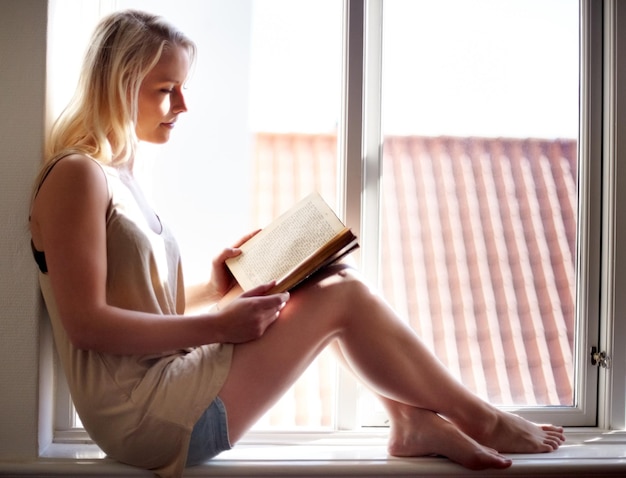 Absorto en su novela Una mujer joven que se relaja en el alféizar de una ventana y lee una novela absorbente