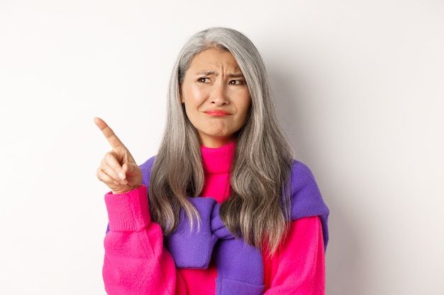 Abuela asiática disgustada en suéter rosa, haciendo muecas y frunciendo el ceño escéptico, señalando la esquina superior izquierda del dedo, de pie sobre fondo blanco.