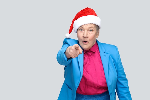 Abuela asombrada en estilo casual colorido, traje azul y gorra roja navideña de pie y mirando y señalando con el dedo a la cámara con cara de sorpresa. interior, tiro del estudio, aislado en fondo gris