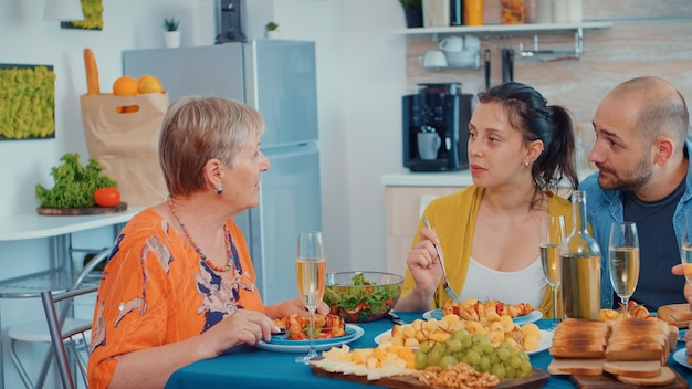 Abuela chasqueando y discutiendo durante la cena. Varias generaciones, cuatro personas, dos parejas felices hablando y comiendo durante una comida gourmet, disfrutando del tiempo en casa, en la cocina sentado junto a la ta