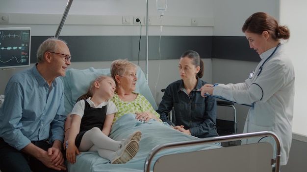 Abuela enferma y familia hablando con el médico sobre la atención médica en la sala del hospital. Especialista médico que explica la enfermedad a un paciente mayor en la cama mientras recibe visitas en la clínica. Visita de chequeo