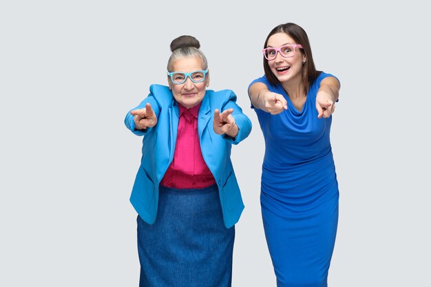 Abuela feliz con nieto señalando con el dedo a la cámara y sonriendo. Relaciones en familia. Relación de nieta o hija con abuela. Foto de estudio de interior, aislado sobre fondo gris