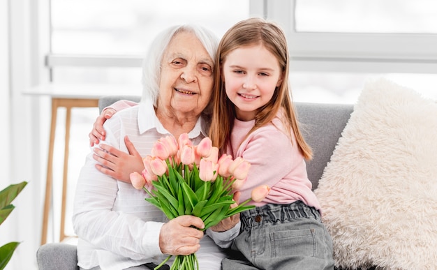 Abuela con flores de tulipanes en sus manos sentada en el sofá con niño nieta y abrazándose