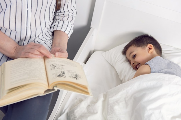 Abuela con gafas y una camisa blanca lee un libro a su nieto acostado en la cama