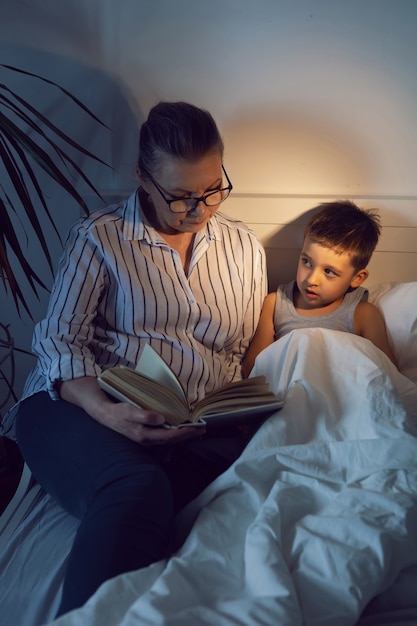 Abuela con gafas y una camisa blanca lee un libro a su nieto acostado en la cama