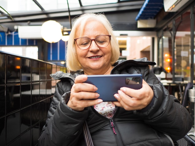 abuela jugadora rubia con gafas jugando en el móvil en la terraza de un restaurante