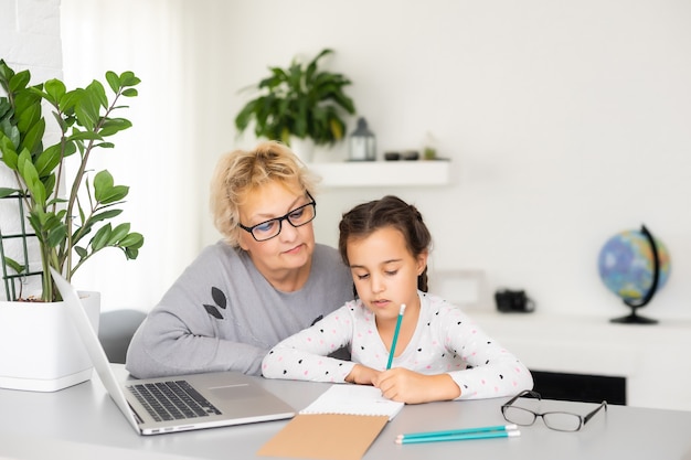 Abuela madura ayudando al niño con la tarea en casa. Abuela satisfecha ayudando a su nieta a estudiar en la sala de estar. Niña escribiendo en un cuaderno con el profesor senior sentado junto a ella