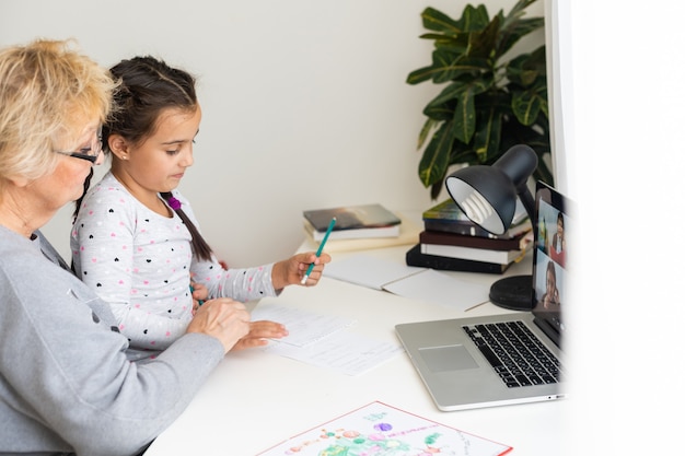 Abuela madura ayudando al niño con la tarea en casa. Abuela satisfecha ayudando a su nieta a estudiar en la sala de estar. Niña escribiendo en un cuaderno con el profesor senior sentado junto a ella