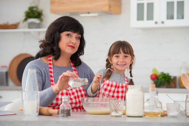 La abuela y la nieta con coletas en delantales de chef cocinan juntas en la cocina batir y salar la mezcla de huevo