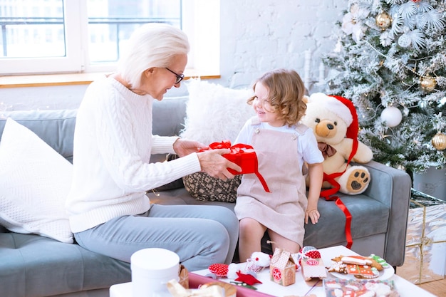 La abuela y la nieta de la familia navideña se sientan en un sofá cerca del árbol de Navidad