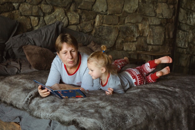 Foto abuela y nieta en pijama de navidad leyendo un libro, acostada en la cama en el chalet.