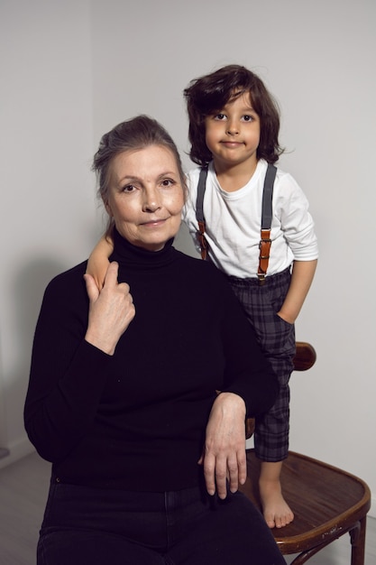 Abuela con un nieto de cuatro años con cabello largo está parado contra una pared blanca con una silla